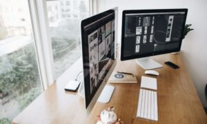escritorio con dos pantallas y un teclado blanco, en el fondo se puede observar una ciudad a través de la ventana