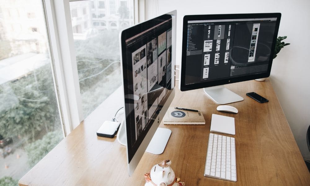 escritorio con dos pantallas y un teclado blanco, en el fondo se puede observar una ciudad a través de la ventana